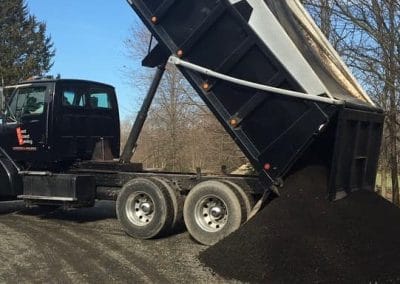 Dump truck unloading dark gravel on a rural road with trees and a small red building nearby.