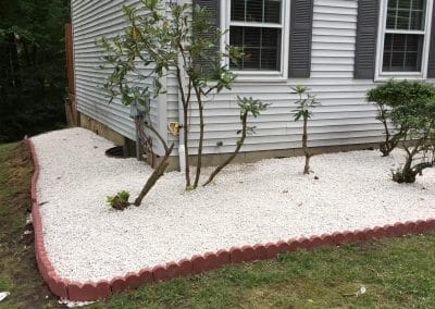 Side of a house with white gravel landscaping and small trees, bordered with red bricks.