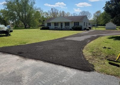 A newly paved driveway leads to a small house surrounded by grass and trees under a blue sky.