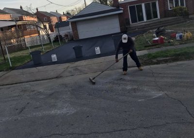Person sweeping a residential street near houses and a driveway with trees in the background.