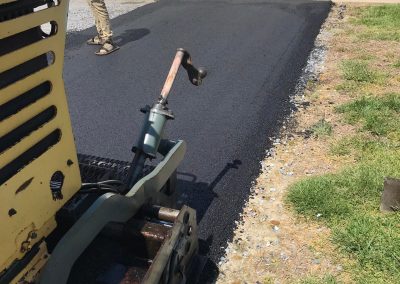 Worker operates paving machine, laying asphalt on a driveway next to a garage.