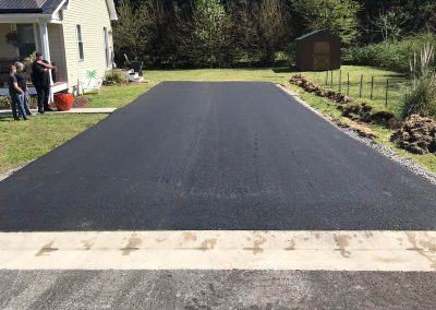 Freshly paved driveway next to a house, with people standing to the left and trees in the background.
