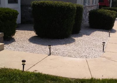 Bushes and white gravel along a house exterior with a yellow tool handle in the foreground.