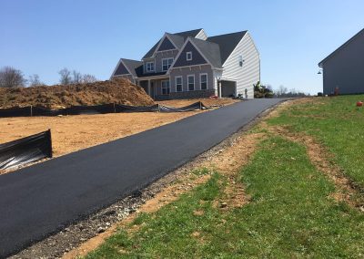Newly paved driveway leading to a large gray house under a clear blue sky. Dirt piles are visible alongside.