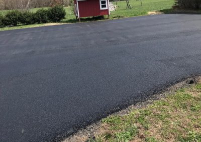 Freshly paved driveway with a small red shed and swing set in the grassy background. Clear blue sky above.