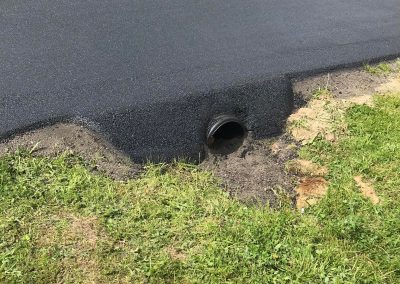 Freshly paved road with a drainage pipe on the edge near a grassy area and a shed in the background.