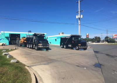 Two black dump trucks parked outside a teal building on a sunny day, with clear skies and visible power lines.