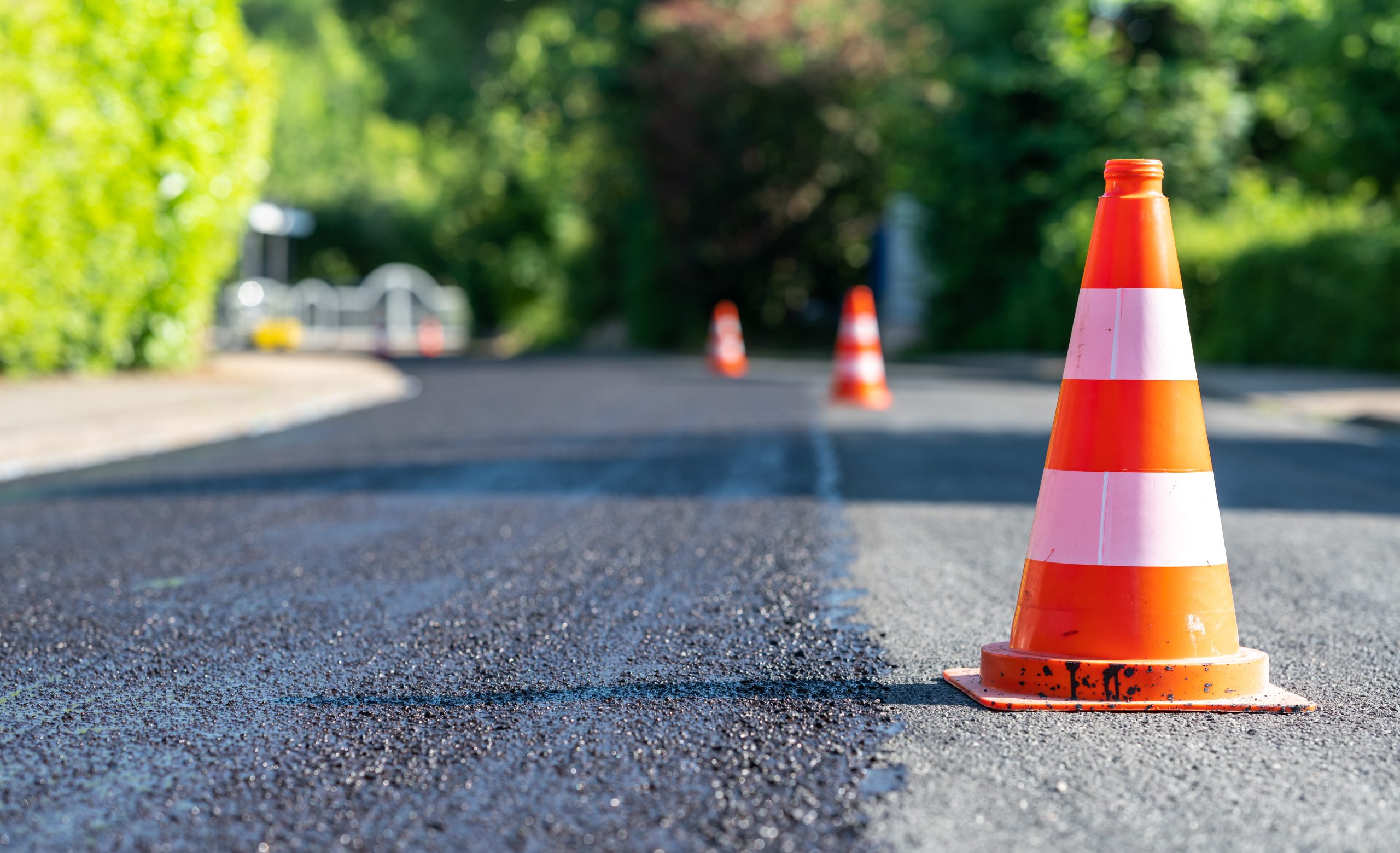 Construction cones marking part of road with a layer of fresh as Orange traffic cones on a freshly paved road with greenery in the background.
