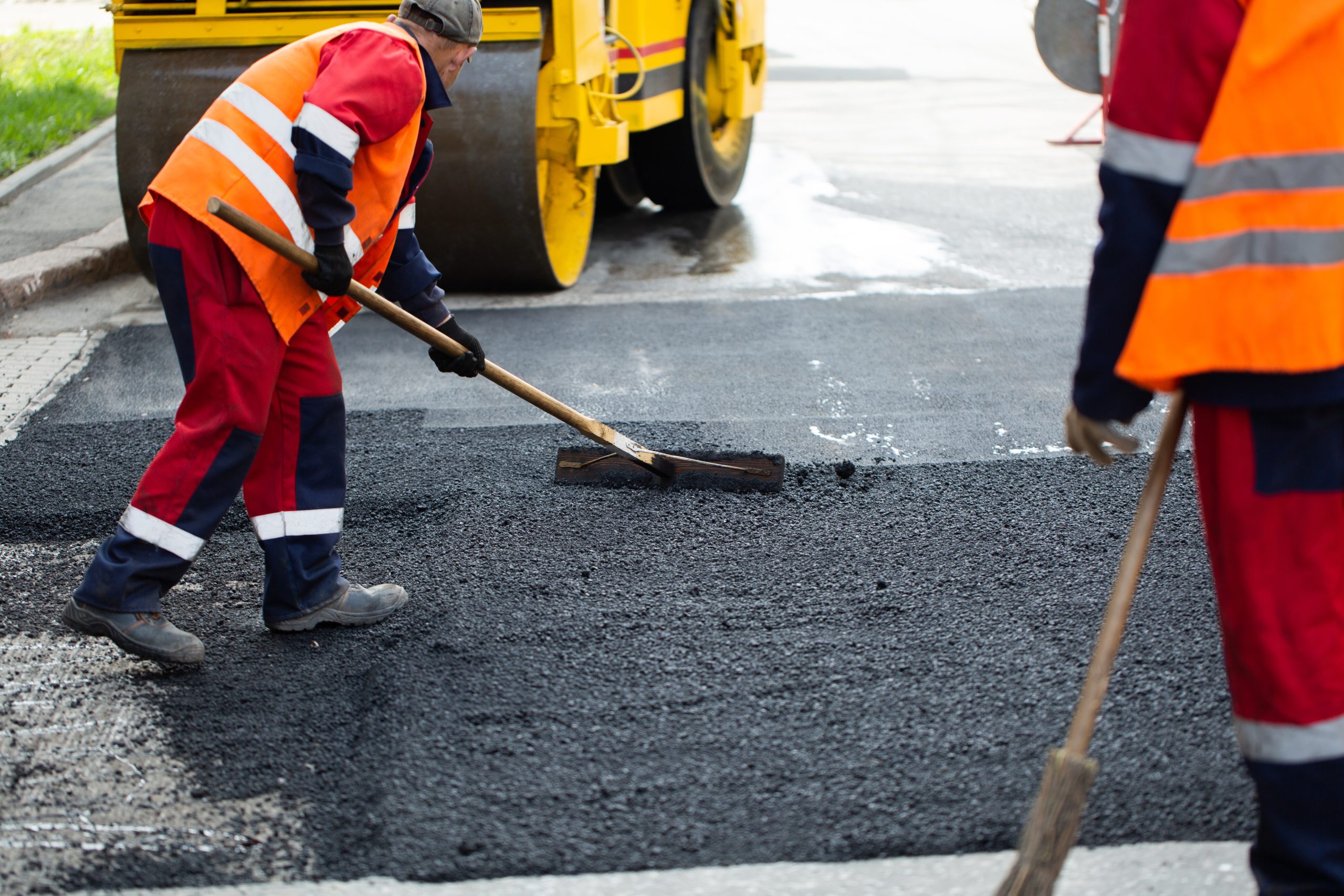 The road workers’ working group updates part of the road with fr Workers in orange vests smooth asphalt on a road, with machinery in the background.
