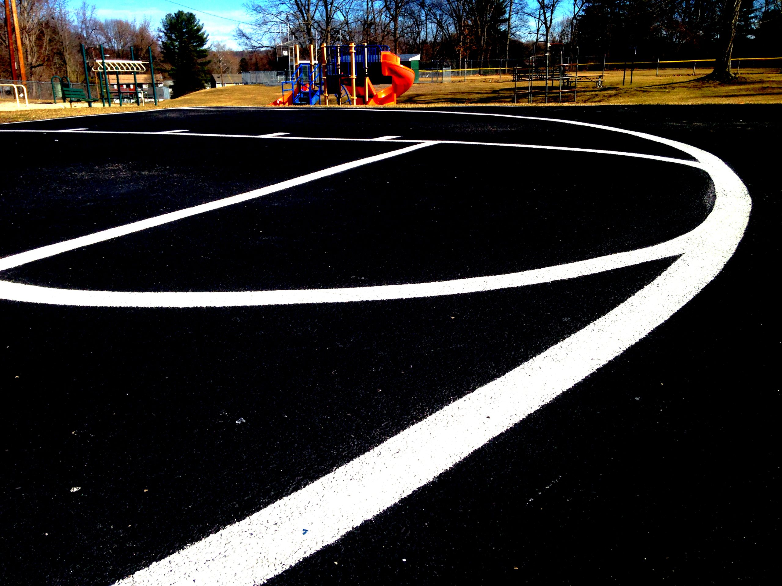 high-angle-view-basketball-court White basketball court lines on blacktop with a playground and trees in the background on a sunny day.
