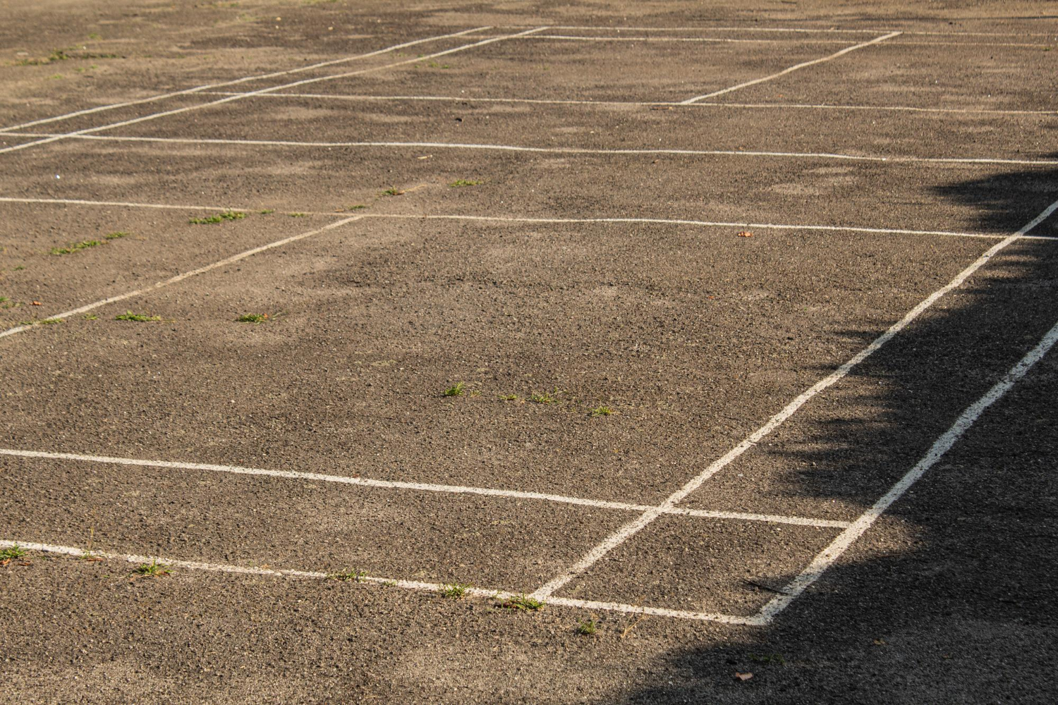 246 Empty asphalt surface with faded white court lines and some grass growing through cracks.