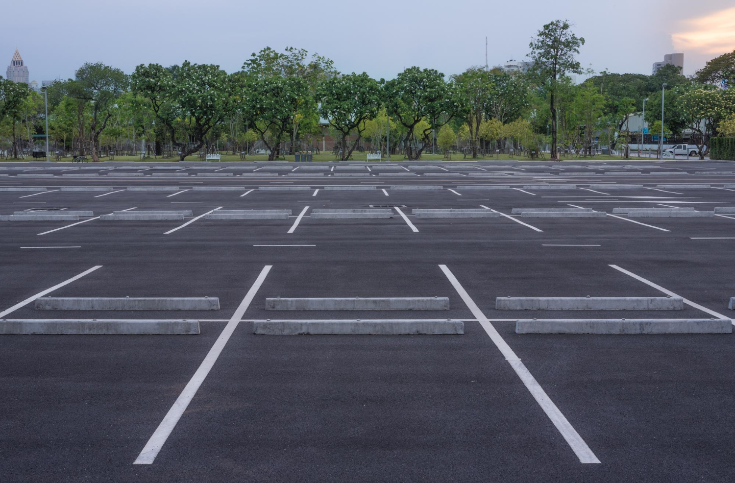 Empty outdoor parking lot with white lines and concrete barriers, trees and greenery in the background.