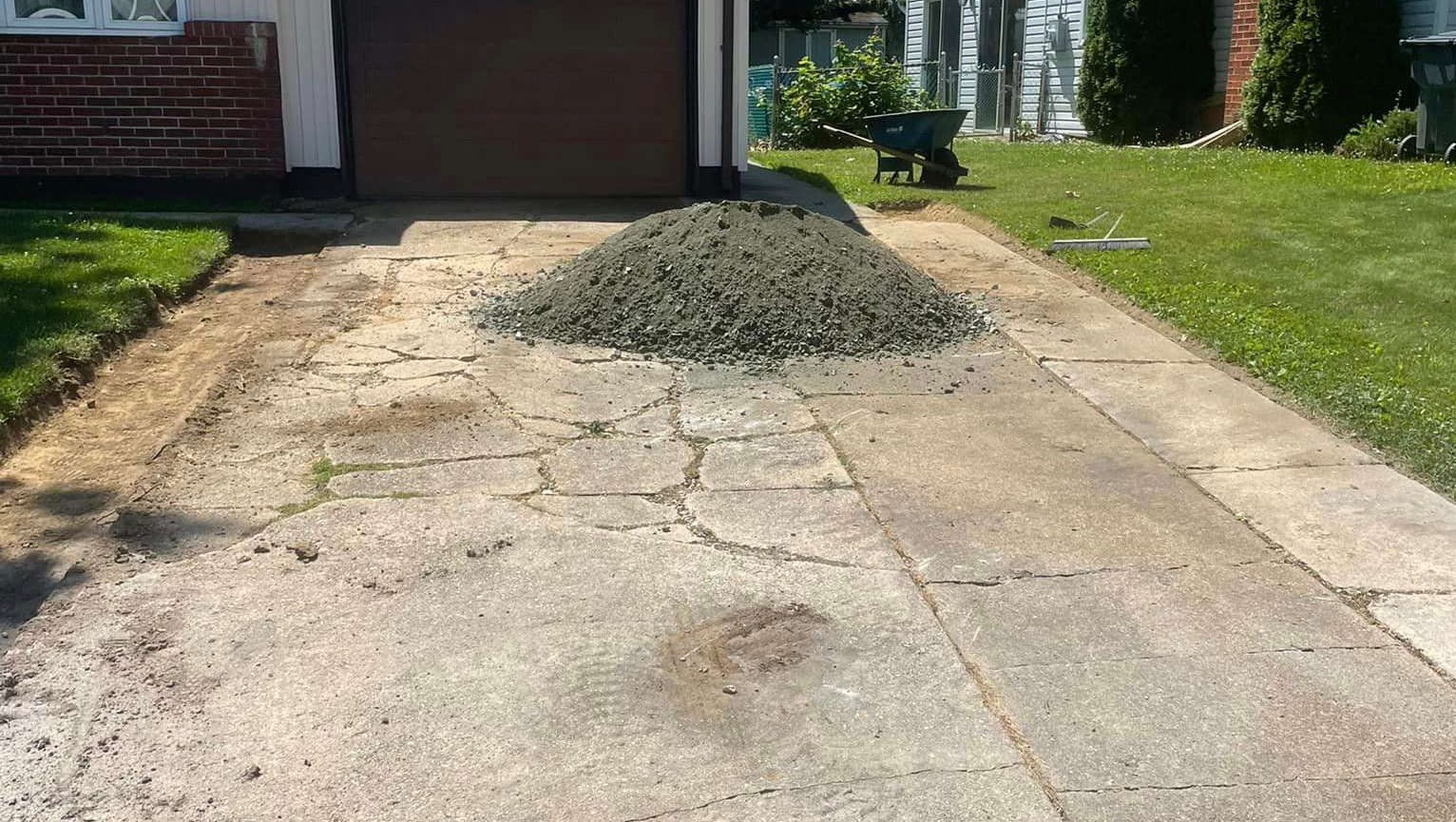 A pile of gravel sits on a cracked driveway in front of a garage on a sunny day.