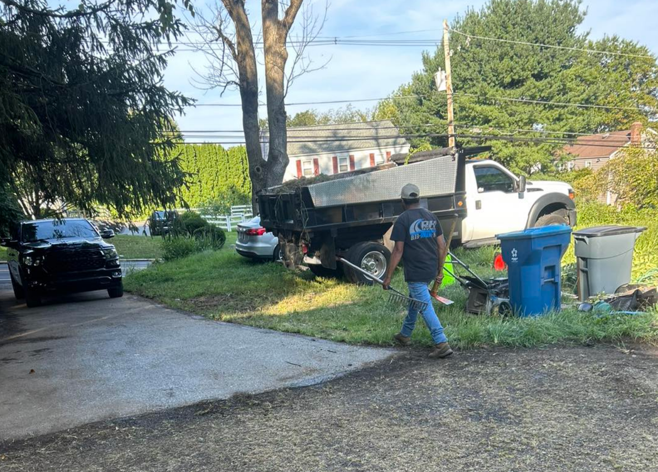 A worker carrying tools walks near a dump truck parked on grass beside blue recycling bins and a driveway.